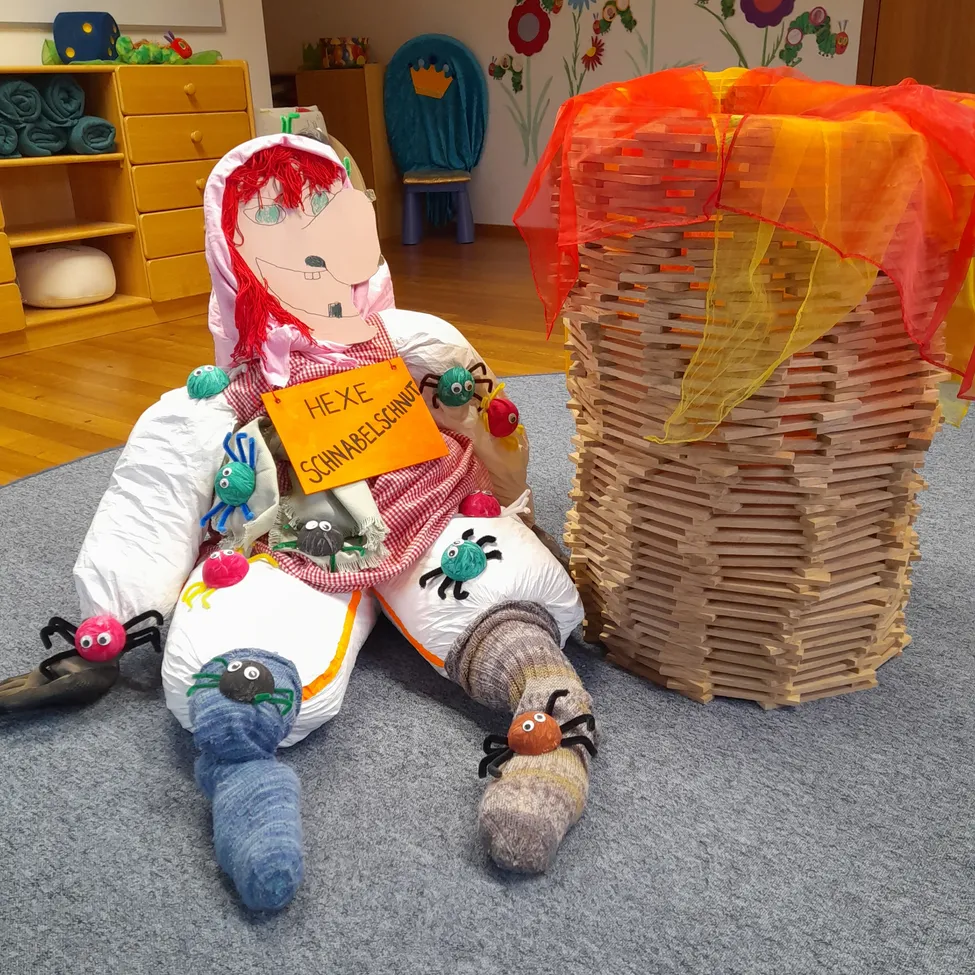 Eine Puppe mit roten Haaren sitzt neben einer Holzkonstruktion mit bunter Abdeckung auf dem Boden. Die Puppe hat ein Schild mit der Aufschrift 'Hexe Schnabbelschnautz'. Spinnen sitzen auf der Puppe und der Holzkonstruktion.