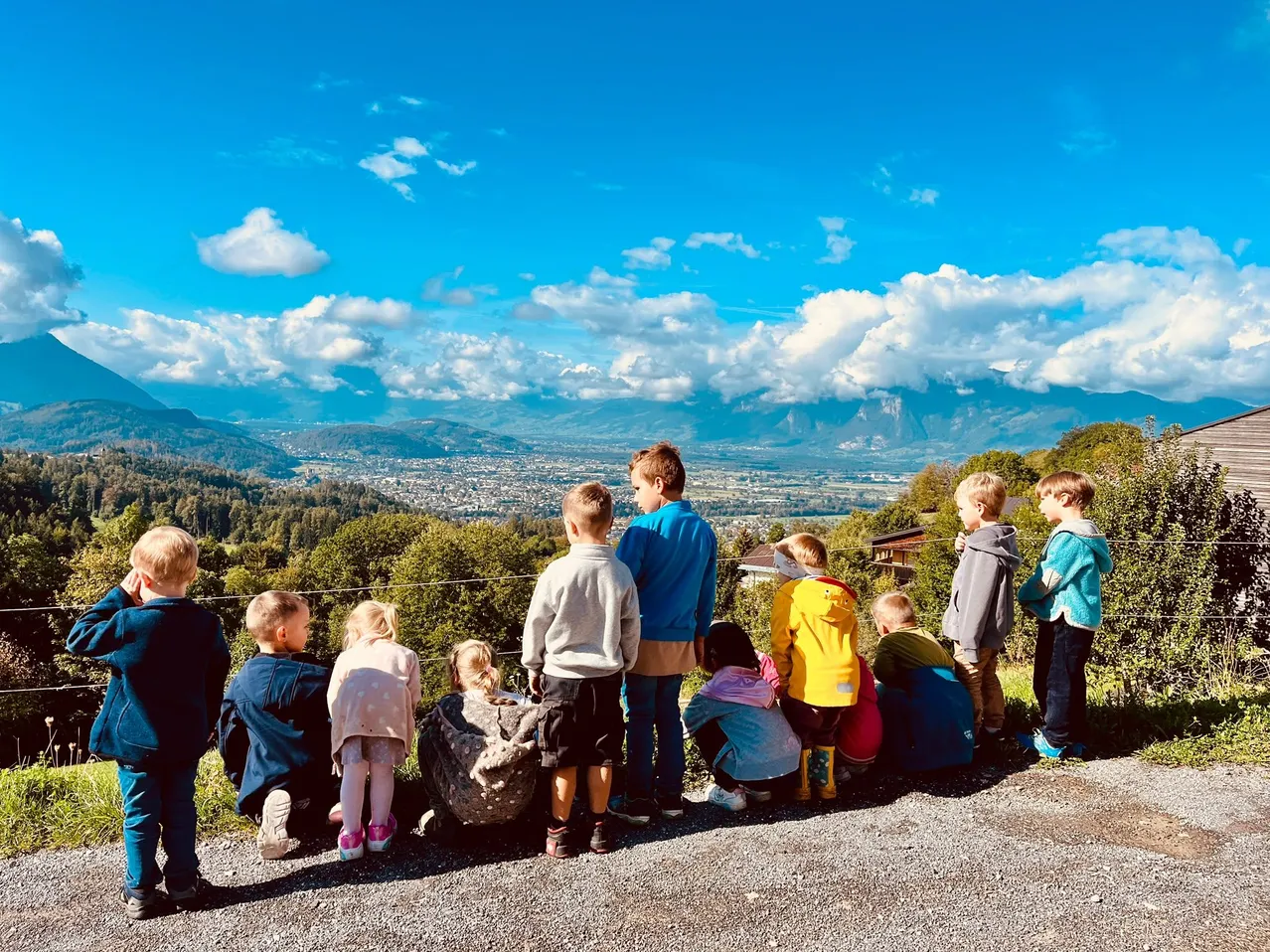 Eine Gruppe von Kindern, einige stehend und einige sitzend, betrachten eine malerische Aussicht auf Berge und eine Stadt unter einem blauen Himmel mit flauschigen Wolken.