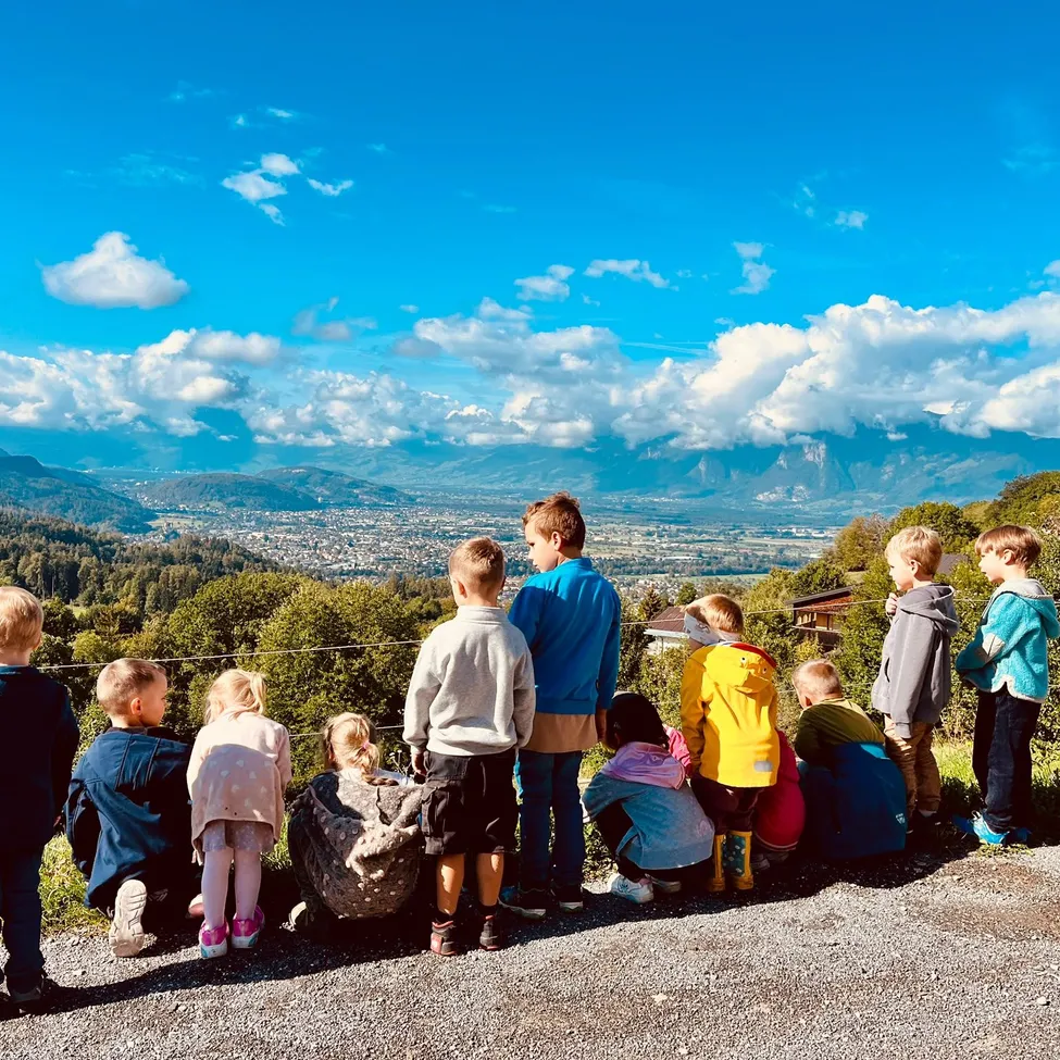 Eine Gruppe von Kindern, einige stehend und einige sitzend, betrachten eine malerische Aussicht auf Berge und eine Stadt unter einem blauen Himmel mit flauschigen Wolken.
