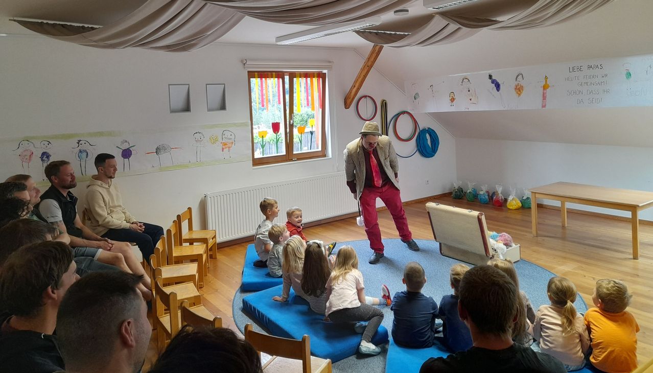 A man entertains a group of children sitting in a circle on blue cushions in a room with white walls and drawings on them.