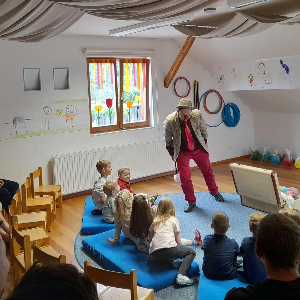 Ein Mann unterhält eine Gruppe von Kindern, die auf blauen Kissen in einem Kreis in einem Raum mit weißen Wänden und Zeichnungen an ihnen sitzen.