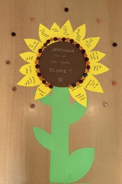 Eine aus Papier gefertigte Sonnenblume mit gelben Blütenblättern und einem braunen Zentrum. Die Blütenblätter haben geschriebene Nachrichten darauf.