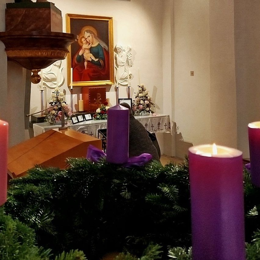 Ein Altar in einer Kirche mit einem religiösen Gemälde, brennenden lila Kerzen und Blumenarrangements auf einem Tisch. Eine Statue und eine Kanzel sind ebenfalls zu sehen.