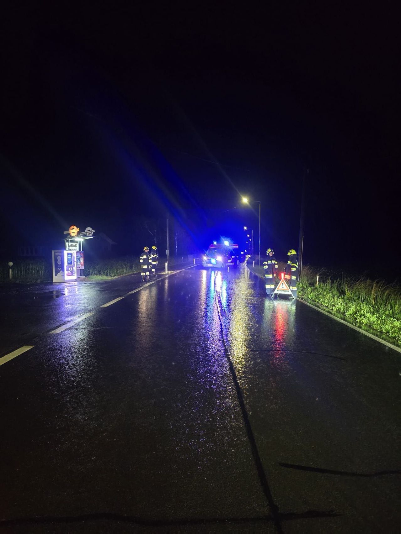 In der Nacht reagieren Rettungsdienste auf einen Vorfall auf einer nassen Straße, wobei blinkende Lichter die Szene beleuchten.