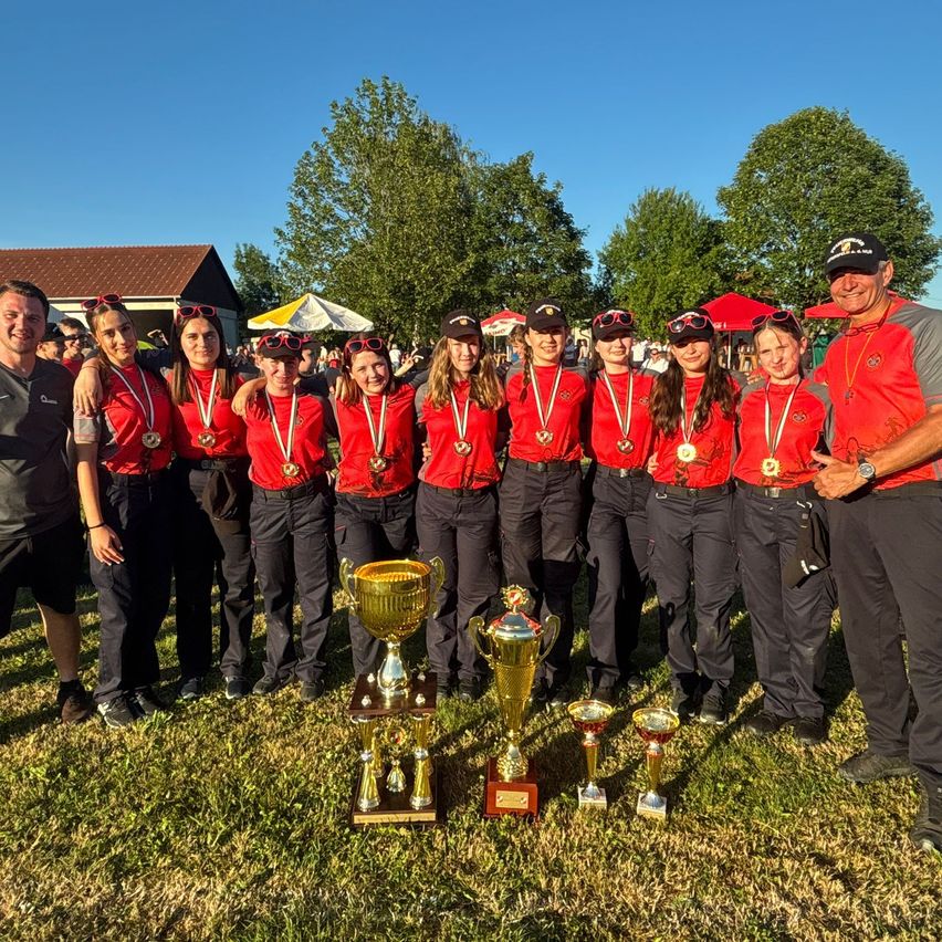 Eine Gruppe von Personen in roten Uniformen posiert mit Trophäen auf einem Grasfeld mit Bäumen im Hintergrund.