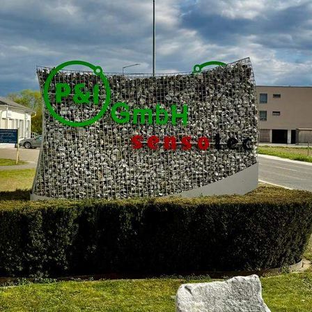 Der Eingang zu einem Gewerbekomplex ist durch eine Steinmauer gekennzeichnet, auf der ein Schild steht, das 'B&S' und 'Sensco' besagt.