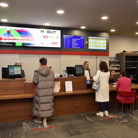 Menschen warten an einer Ticketschalter in einem Bahnhof. Monitore zeigen Informationen über dem Schalter an. Eine Frau in einem weißen Mantel steht neben einem Stuhl.