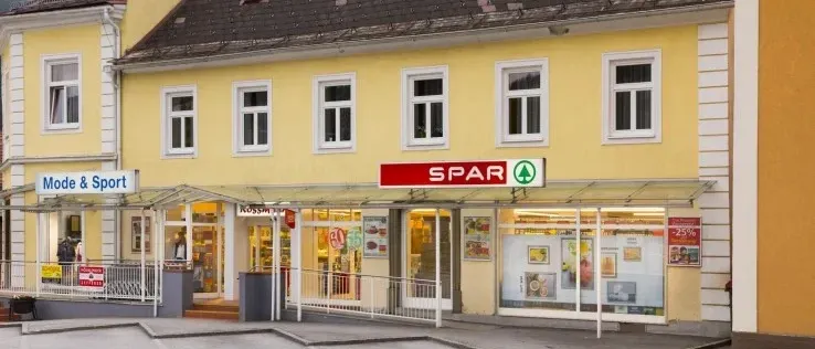 Ein gelbes Gebäude mit einem schwarzen Dach und einem roten Schild, das Spar steht. Das Gebäude hat mehrere Fenster und einen Glaseingang. Vor dem Gebäude befindet sich ein Vordach.