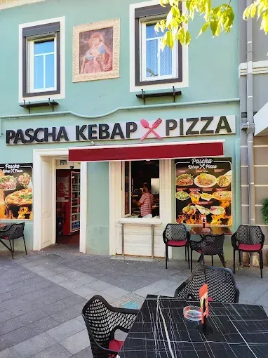Ein Restaurant namens Pascha Kebab Pizza mit Tischen, Stühlen und einer Person im Inneren. Das Restaurant hat eine grüne Wand mit einem Bilderrahmen.