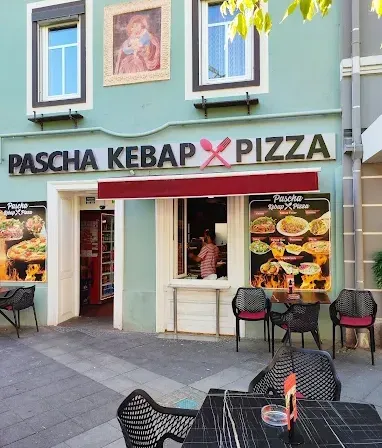 Ein Restaurant namens Pascha Kebab Pizza mit Tischen, Stühlen und einer Person im Inneren. Das Restaurant hat eine grüne Wand mit einem Bilderrahmen.