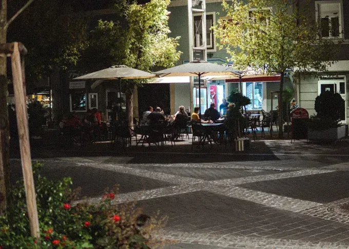 Ein Nachtblick auf einen Stadtplatz mit Menschen, die um Tische unter Schirmen vor einem Restaurant sitzen.