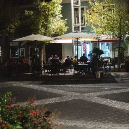 Ein Nachtblick auf einen Stadtplatz mit Menschen, die um Tische unter Schirmen vor einem Restaurant sitzen.