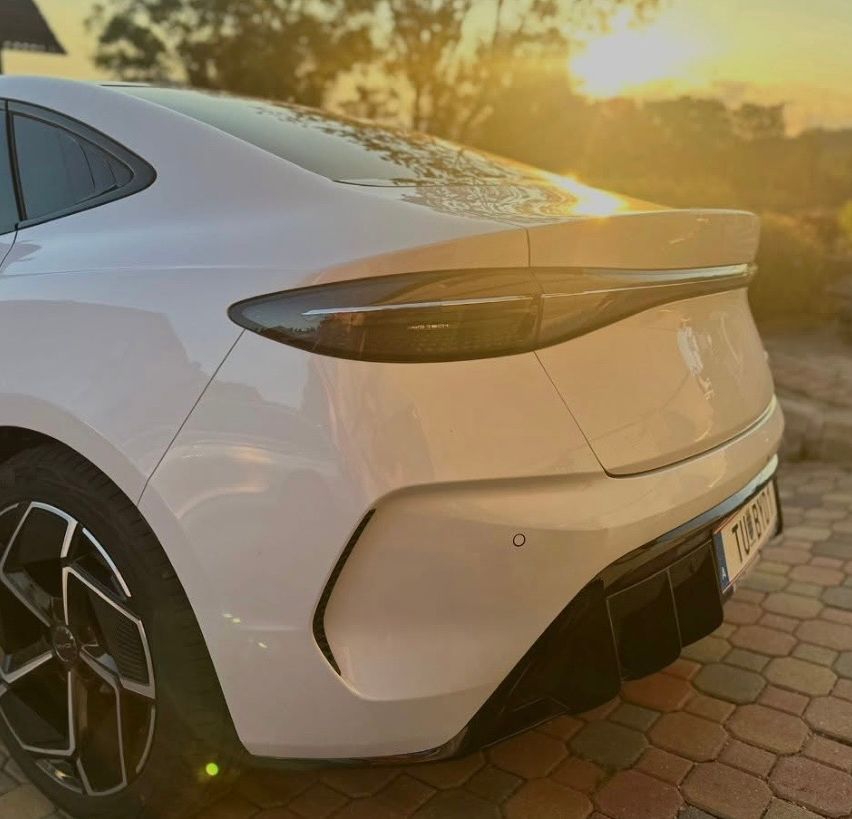 Ein weißer Wagen steht auf einem gepflasterten Weg bei Sonnenuntergang. Der Heck des Autos hat einen Spoiler und ein Nummernschild. Die Scheinwerfer sind an.