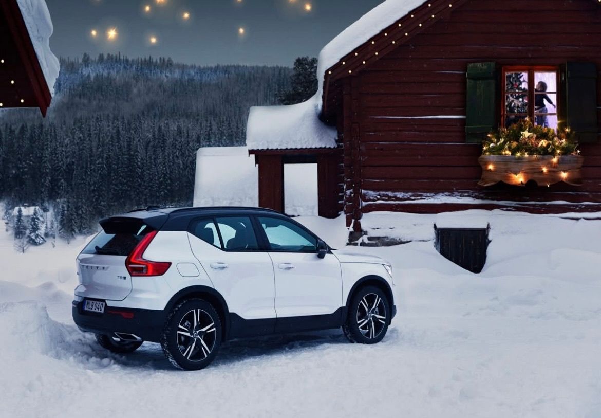 Ein weißer SUV, der im Winter vor einer Holzhütte geparkt ist, mit Schnee, Sternen am Himmel und dekorativen Lichtern auf dem Dach.