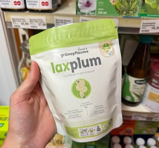 Eine Hand hält einen Beutel mit grünen Pflaumen, der laxplum für die Verdauungsgesundheit kennzeichnet, in einem Geschäft mit verschiedenen Produkten auf den Regalen.