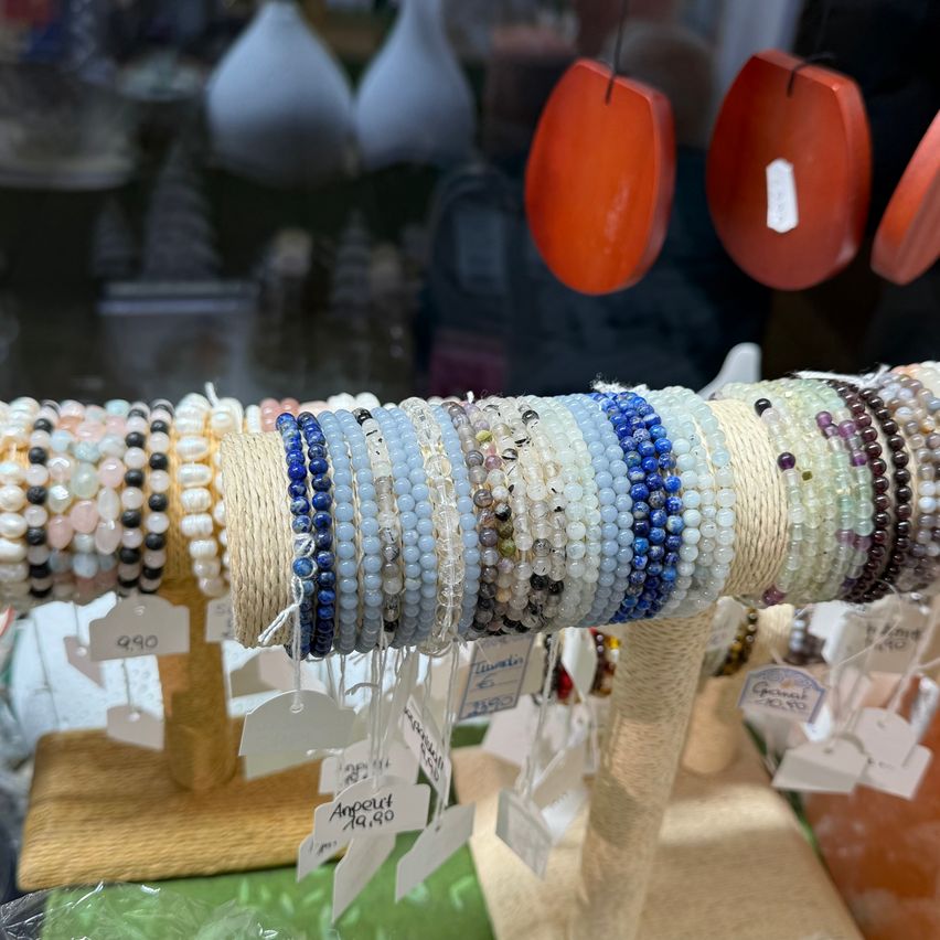Eine Ausstellung von Perlenarmbändern ist auf einem Holzständer in einem Geschäft angeordnet. Die Armbänder bestehen aus verschiedenen Materialien, einschließlich blauer und weißer Steine, und hängen an einer Schnur. Preisschilder sind an jedem Armband befestigt. Hinter den Armbändern befinden sich mehrere andere ausgestellte Gegenstände, einschließlich orangefarbener Objekte.