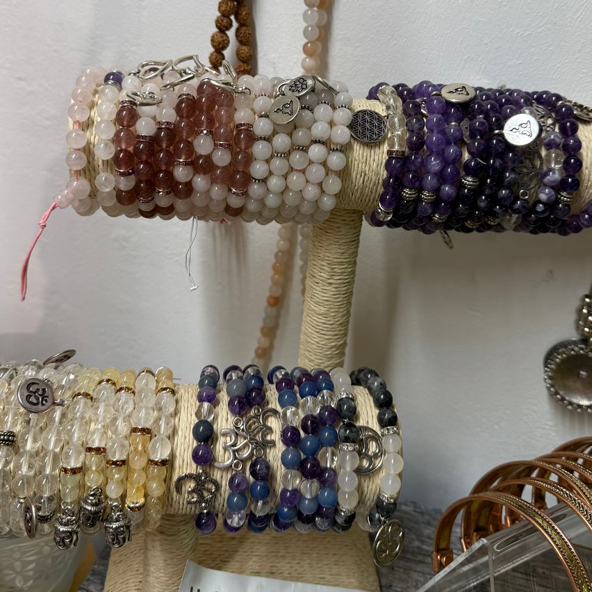 Verschiedene Perlenarmbänder sind auf einem Holzständer ausgestellt. Die Armbänder bestehen aus verschiedenfarbigen Perlen und haben silberne Anhänger. Der Ständer besteht aus Seil und hat einen weißen Hintergrund.