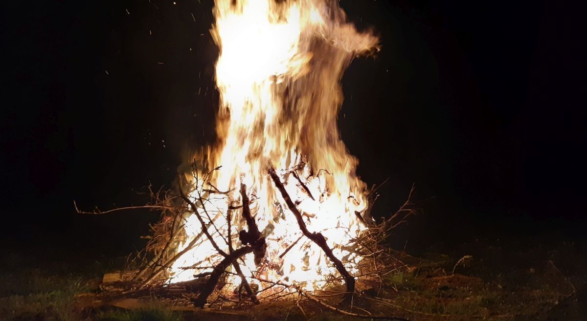 Ein Lagerfeuer brennt hell in der Nacht, umgeben von einem Ring aus Holz und Ästen, der ein warmes Licht spendet.