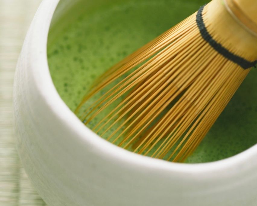 Ein Schneebesen mit Bambussprossen sitzt in einer weißen Schüssel und mischt eine leuchtend grüne Flüssigkeit. Der Griff des Schneebesens ist schwarz.