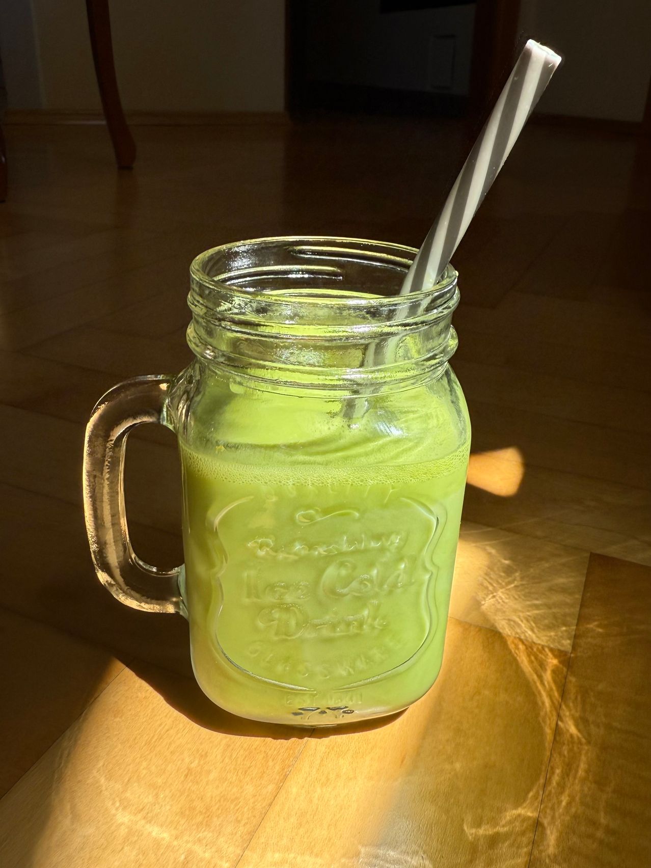 Ein grüner Smoothie in einem Einmachglas mit einem gestreiften Strohhalm, auf einem Holztisch stehend. Das Sonnenlicht scheint auf den Tisch.