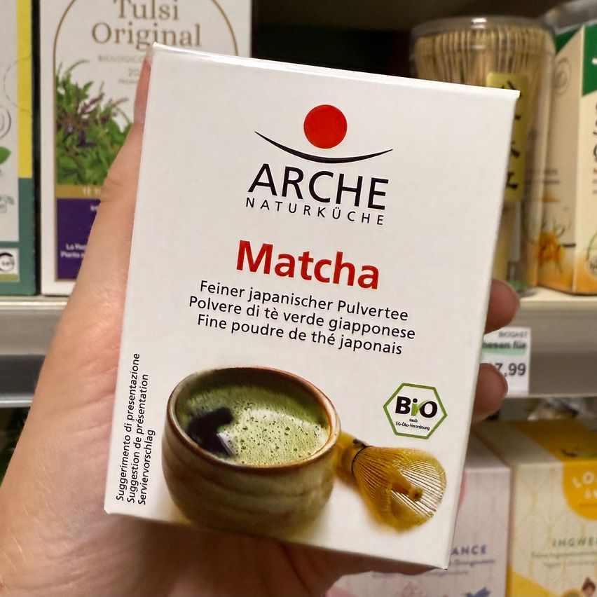 Hand hält eine Schachtel Arche Naturküche Matcha. Die Verpackung zeigt ein grünes Teeblatt und einen Besen und ist weiß und rot.