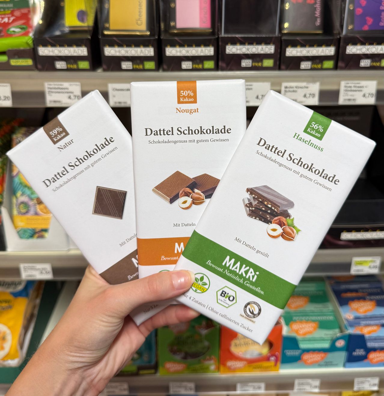 Eine Hand hält drei Schokoladenschachteln in einem Geschäft, beschriftet mit 'Dattel Schokolade' und 'Makri', mit verschiedenen Kakaoprozenten und Zutaten.