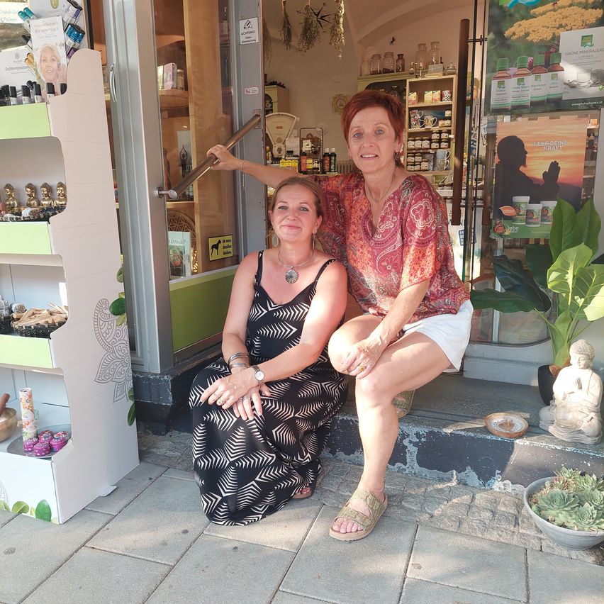 Zwei Frauen sitzen vor einem Geschäft, eine trägt ein schwarzes Kleid und die andere ein rotes Oberteil mit weißen Shorts.