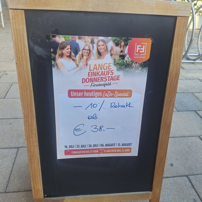 Ein hölzernes Schild zeigt ein Plakat für einen langen Einkaufstag in Furstenfeld. Das Plakat zeigt drei Frauen mit Getränken und einem Logo. Das Sonderangebot umfasst Rabatte auf Robatsch und E 38. Die Veranstaltung läuft vom 16. Juli bis 13. August.