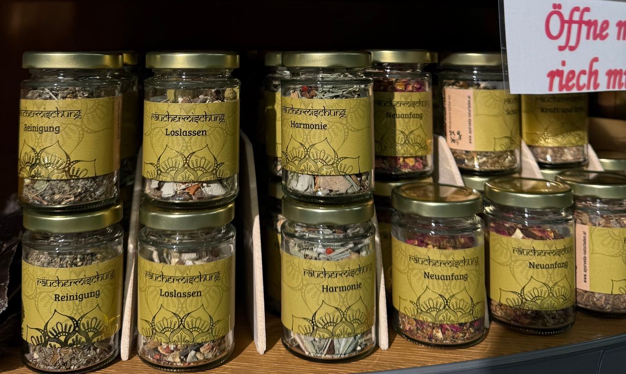 Eine Sammlung von Glasgläsern mit goldenen Deckeln, die jeweils eine andere Mischung aus Kräutern und Blumen enthalten, beschriftet mit 'Loslassen' und 'Neuanfang'. Die Gläser sind auf einem Holzregal gestapelt.
