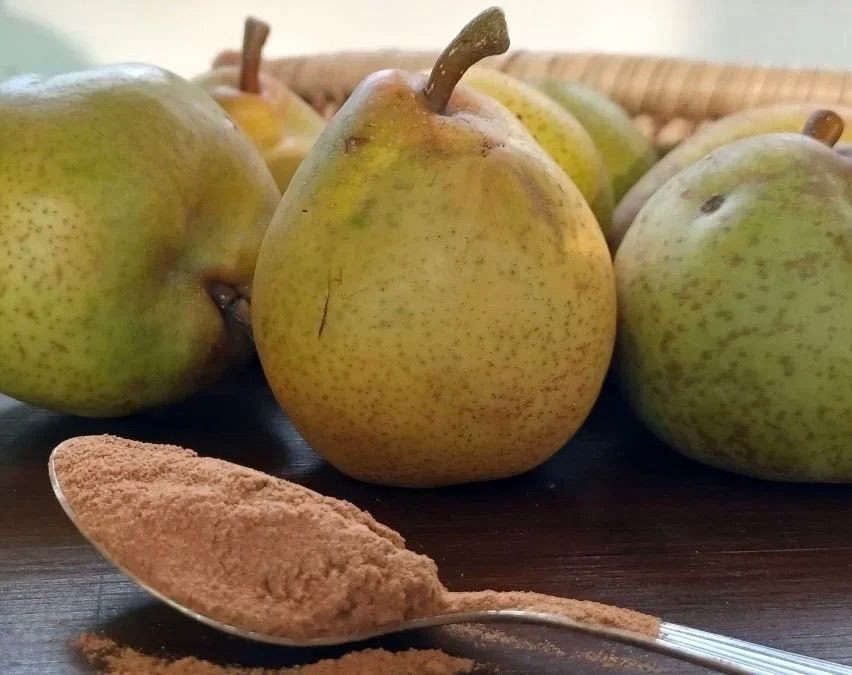A close-up of pears with a spoon of cinnamon powder on a wooden table. The pears are green and yellow with some brown spots.