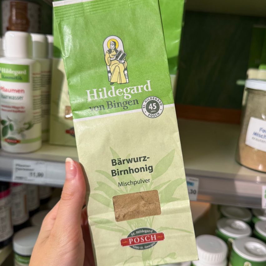A hand holds a green bag of Hildegard von Bingen's Barwurz-Birnhonig mix powder in a store. The bag is labeled with a drawing of a woman and the brand logo.