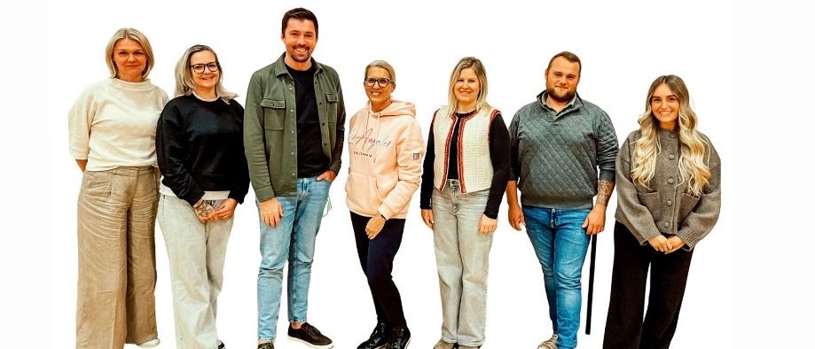 Eine Gruppe von Menschen, darunter drei Frauen und drei Männer, steht eng beieinander und lächelt, gekleidet in lässige Winterkleidung.
