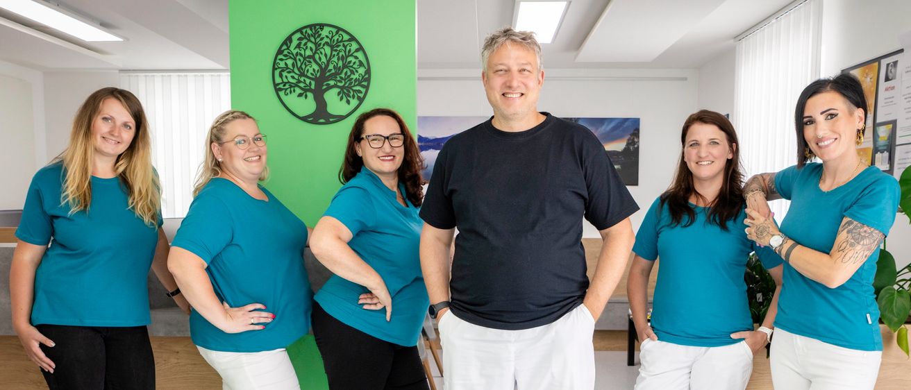 Fünf Personen in passenden blauen Shirts und weißen Hosen stehen in einem Raum. Ein Mann in einem schwarzen Shirt und weißen Hosen steht in der Mitte. Hinter ihnen befindet sich eine grüne Säule mit einem Baumdesign.