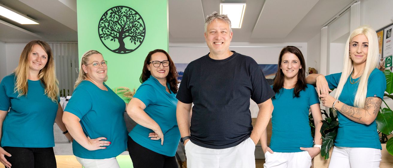 Fünf Personen stehen in einem Raum und tragen passende blaue Shirts. Der Mann in der Mitte trägt ein schwarzes Shirt und weiße Hosen. Die Frau rechts trägt einen Ring. Sie posieren für ein Foto.