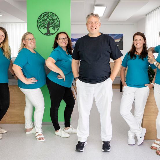 Eine Gruppe von Menschen in passenden blauen Shirts und weißen Hosen steht in einem Büro zusammen. Ein Mann in einem schwarzen Shirt steht lächelnd in der Mitte.
