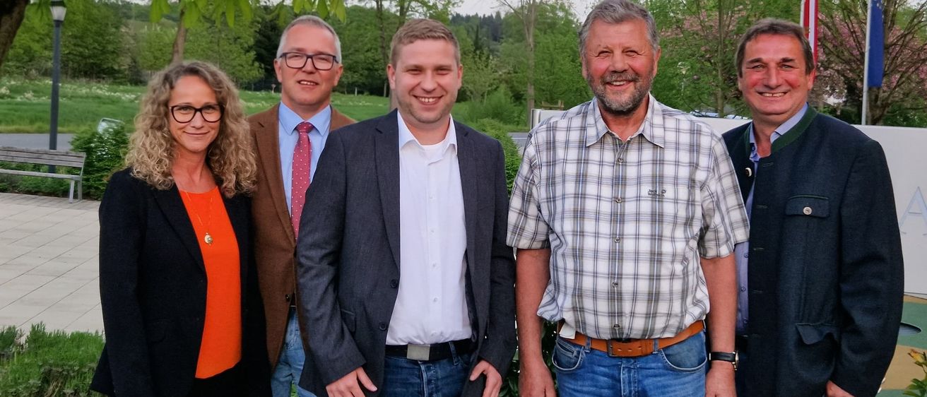Fünf Personen stehen zusammen in einem Garten. Eine Frau links, zwei Männer in der Mitte und zwei weitere Männer rechts. Sie alle lächeln und scheinen für ein Foto zu posieren. Hinter ihnen stehen mehrere Bäume und ein Rasenfeld.