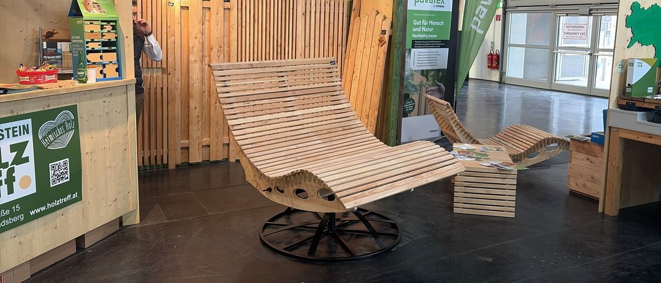 Ein Holzloungestuhl mit Radbasis steht in einem Raum mit Holzwänden zur Schau. Eine Person ist links teilweise sichtbar und ein Banner befindet sich rechts.