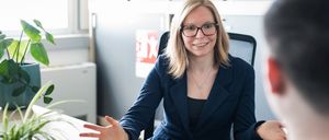 Eine Frau mit Brille und blauer Jacke sitzt in einem Büro an einem Schreibtisch, lächelt und spricht mit jemandem. Sie trägt eine Halskette und ist von roten Karten mit der Nummer '9' umgeben.