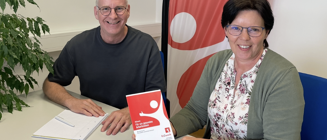 A man and woman sit at a desk with a book titled 'Fit for Work'. They smile and pose for a photo. The man wears glasses and a black shirt, the woman a floral shirt and green cardigan. A notebook and pen are on the table. A flag with red and white design is behind them.