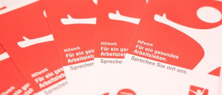 Mehrere rote und weiße Karten mit der Aufschrift 'fit2work'. Die Karten enthalten Text über ein gesundes Arbeitsleben und eine Kontaktnummer.