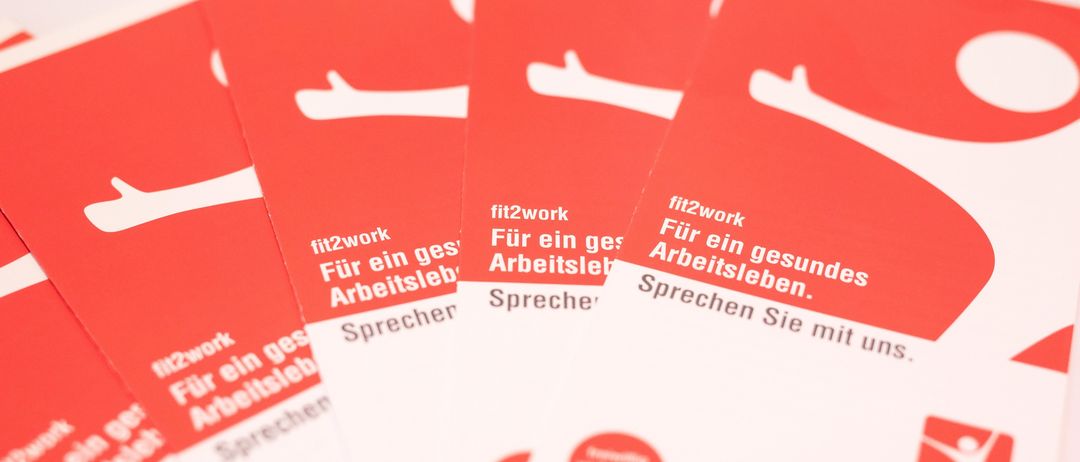 Mehrere rote und weiße Karten mit der Aufschrift 'fit2work'. Die Karten enthalten Text über ein gesundes Arbeitsleben und eine Kontaktnummer.