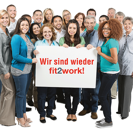 Eine Gruppe von verschiedenen Personen, die lächelnd zusammenstehen und ein weißes Schild mit dem Text 'Wir sind wieder fit2work!' in roten Buchstaben halten.