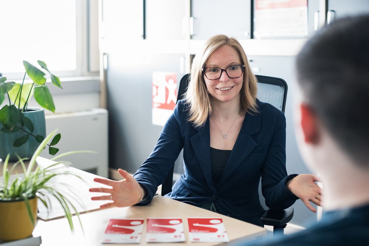 Eine Frau mit Brille sitzt in einem Bürostuhl, lächelt und scheint ein Gespräch zu führen. Auf einem Schreibtisch vor ihr liegen drei Karten mit dem Buchstaben S.