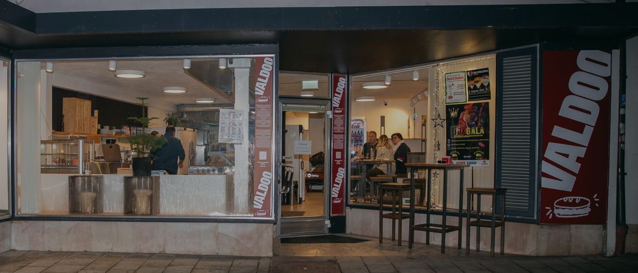 Das Äußere eines Valdoon-Restaurants bei Nacht. Leute sitzen an Tischen im Inneren. Ein Mann steht an der Theke, und ein anderer Mann sitzt in der Nähe.