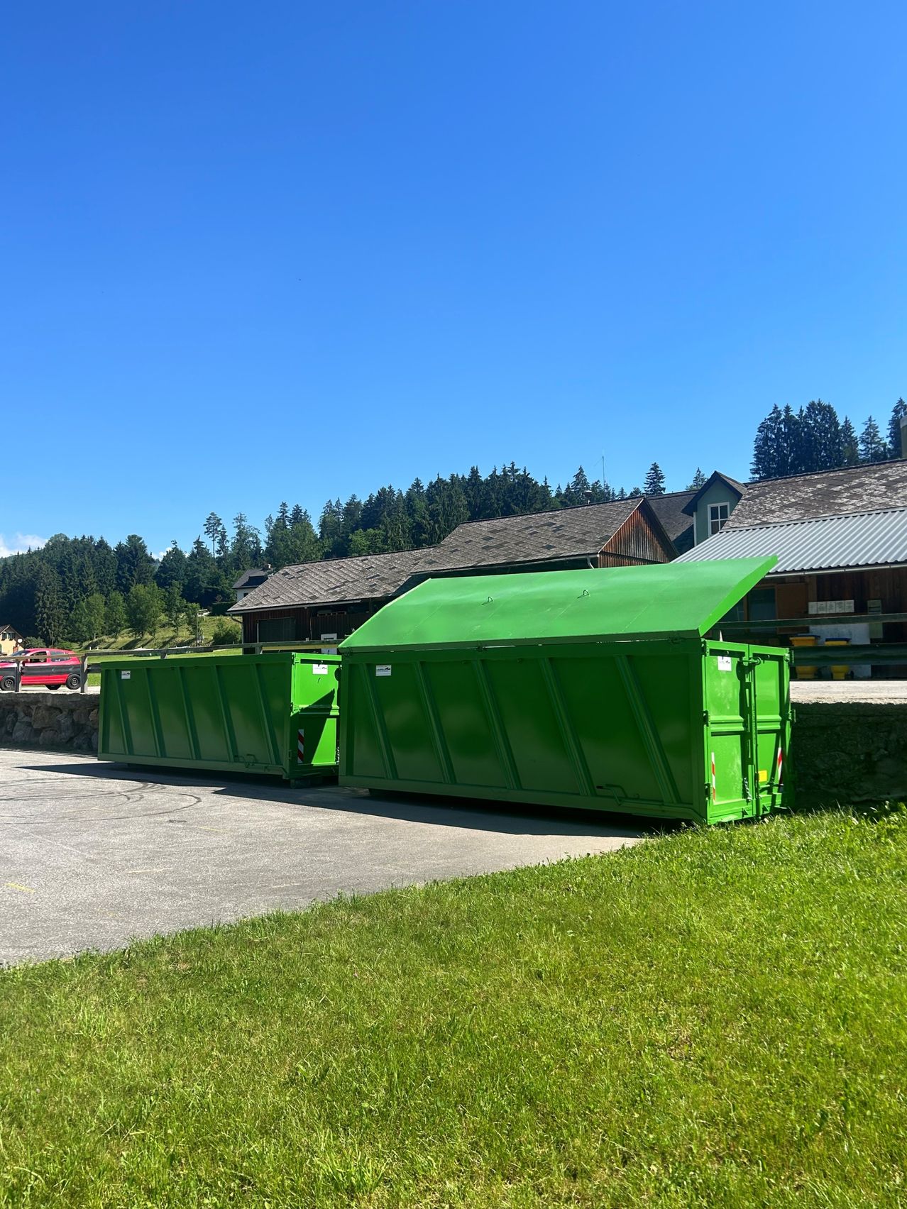 Zwei grüne Müllcontainer stehen im Vordergrund einer ländlichen Straße. Dahinter befindet sich ein Haus mit einem Metalldach. Bäume und Berge sind im Hintergrund zu sehen.