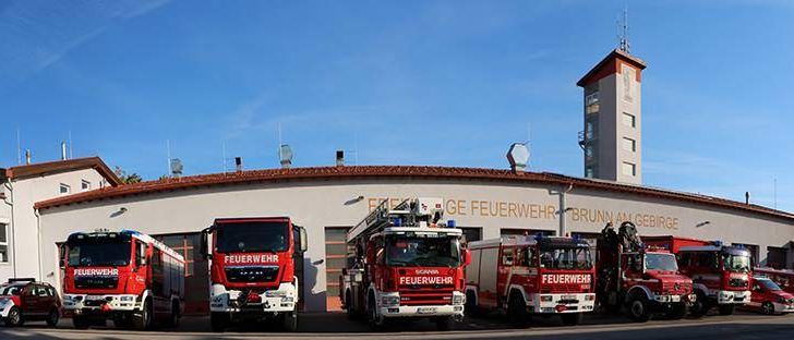 Eine Reihe roter Feuerwehrfahrzeuge steht vor einem weißen Gebäude mit Turm, unter einem blauen Himmel mit einigen Wolken.