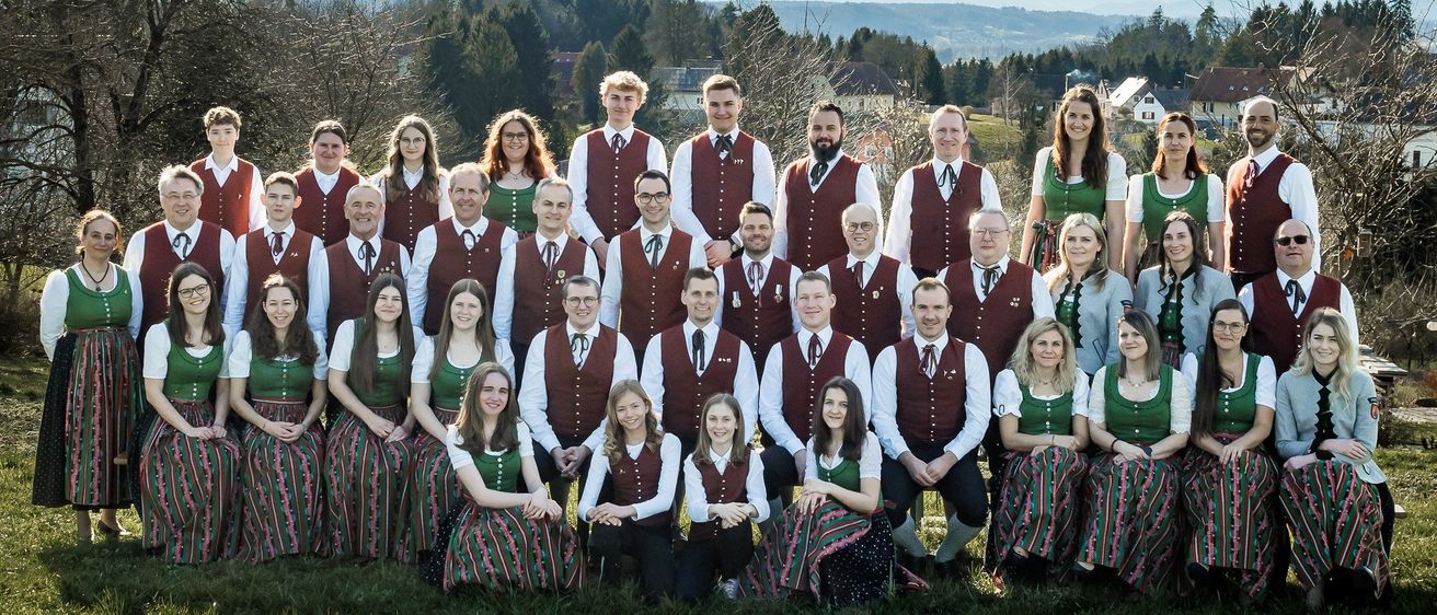 Eine Gruppe von Menschen, die in traditioneller Kleidung mit grünen Westen und roten Krawatten gekleidet sind, posiert für ein Foto in einer Outdoor-Umgebung mit einer Bergkette im Hintergrund.