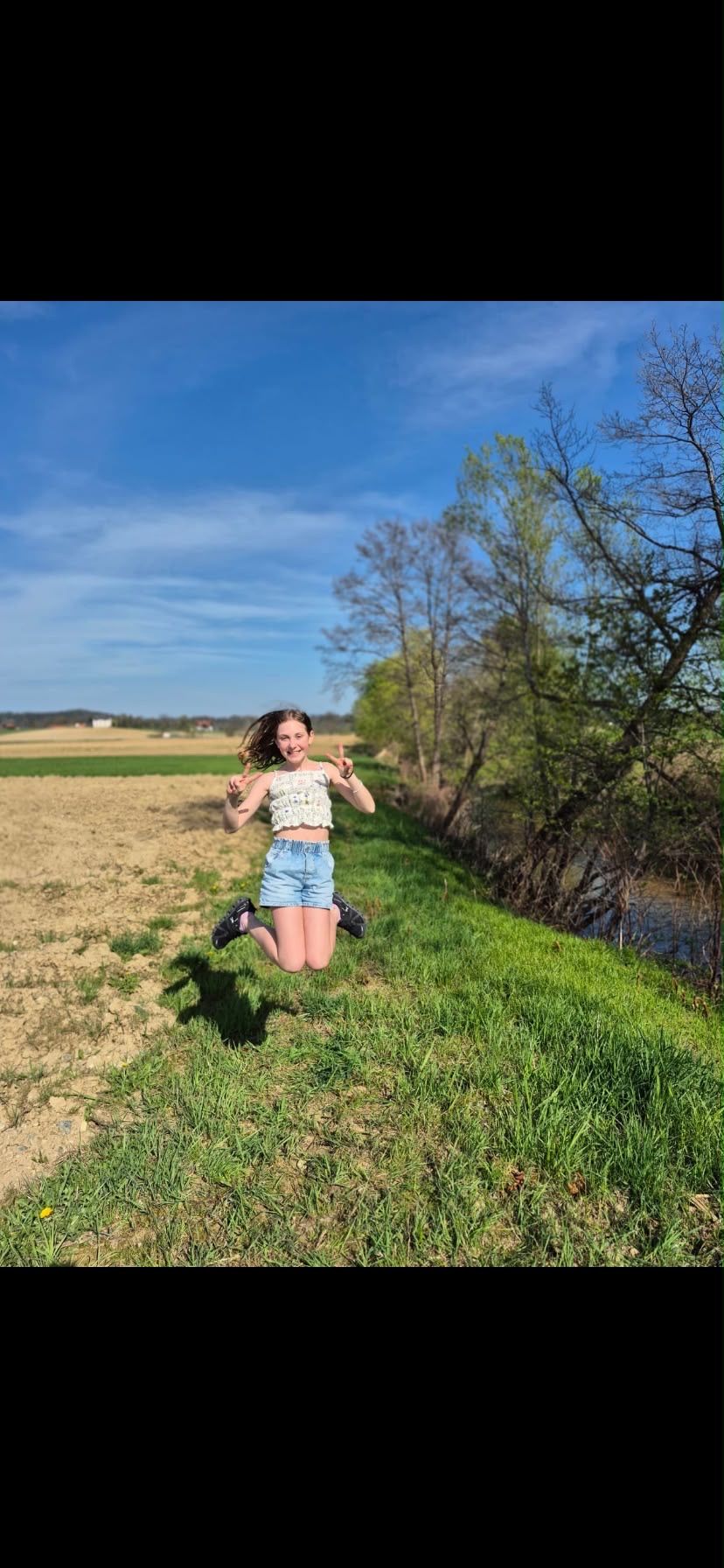 A young woman jumps in the air with a peace sign, standing on a grassy field with trees and a small stream in the background.