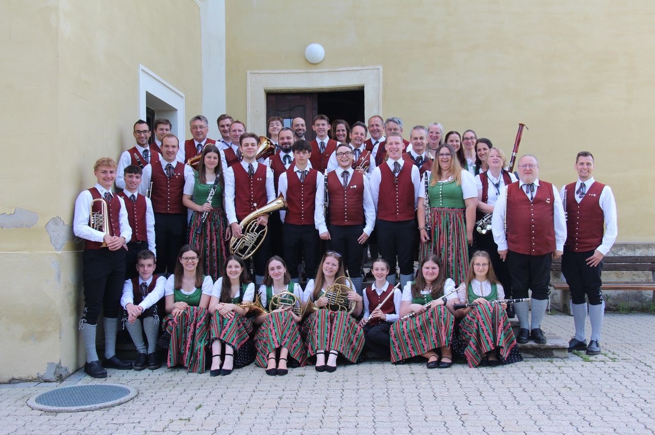 Ein Musikverein in traditioneller Kleidung posiert für ein Foto vor einem Gebäude. Sie halten verschiedene Musikinstrumente in den Händen.