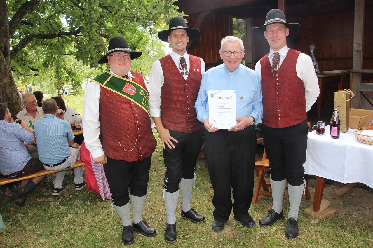 Four men in traditional attire, two wearing hats, one holding a certificate, standing on grass with a wooden structure and trees in the background.